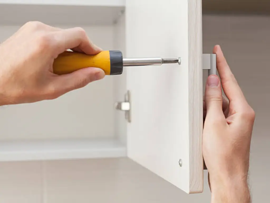 The worker sets a new handle on the white cabinet with a screwdriver